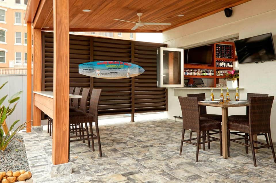 Covered outdoor bar and dining area with wood beams, wicker seating, and surfboard decoration