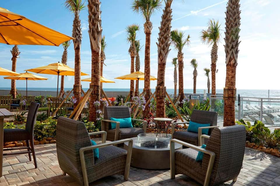 Oceanfront lounge with fire pit, wicker seating, palm trees, and yellow umbrellas