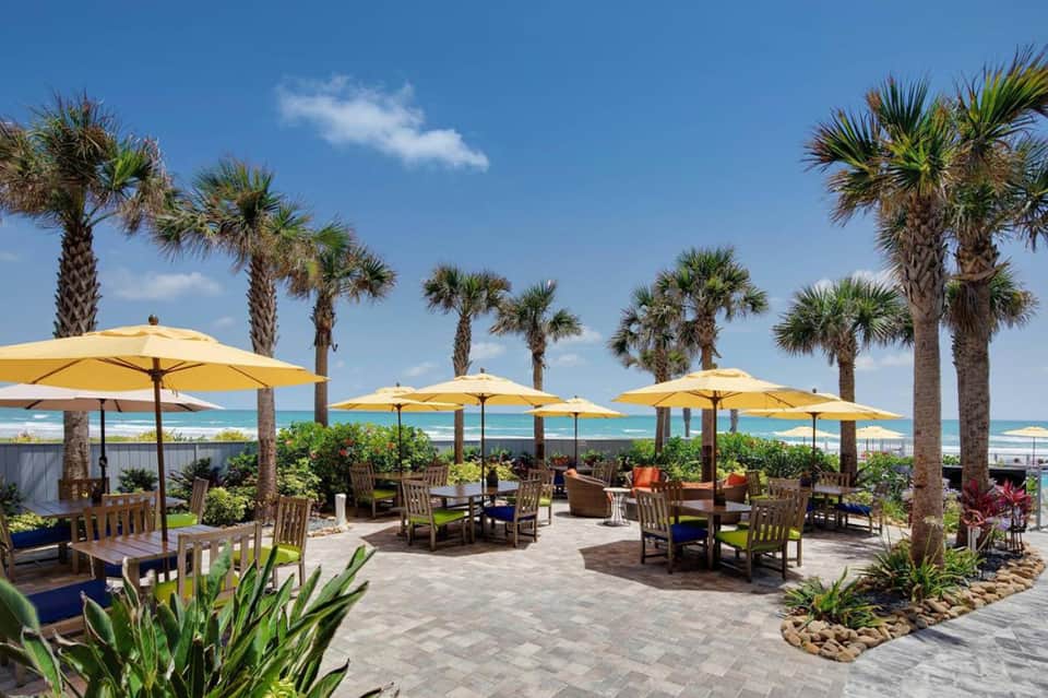 Beachfront resort patio with yellow umbrellas, palm trees, and ocean view seating