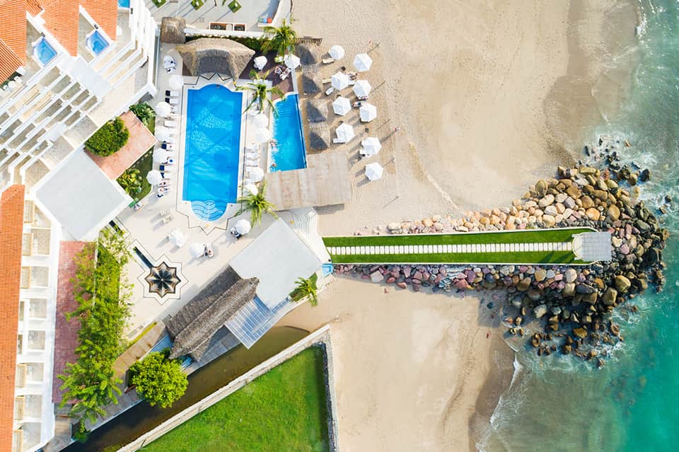 Aerial view of beachfront resort with pools, sandy beach, palm trees, and ocean access