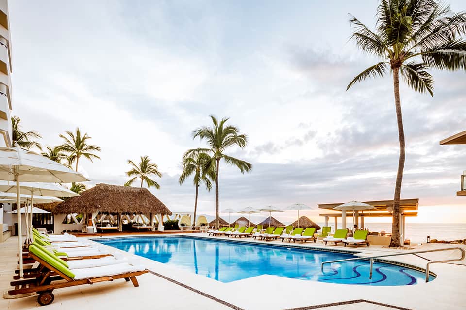 Beachfront resort pool at dusk with palm trees, white loungers, tiki hut, and ocean horizon