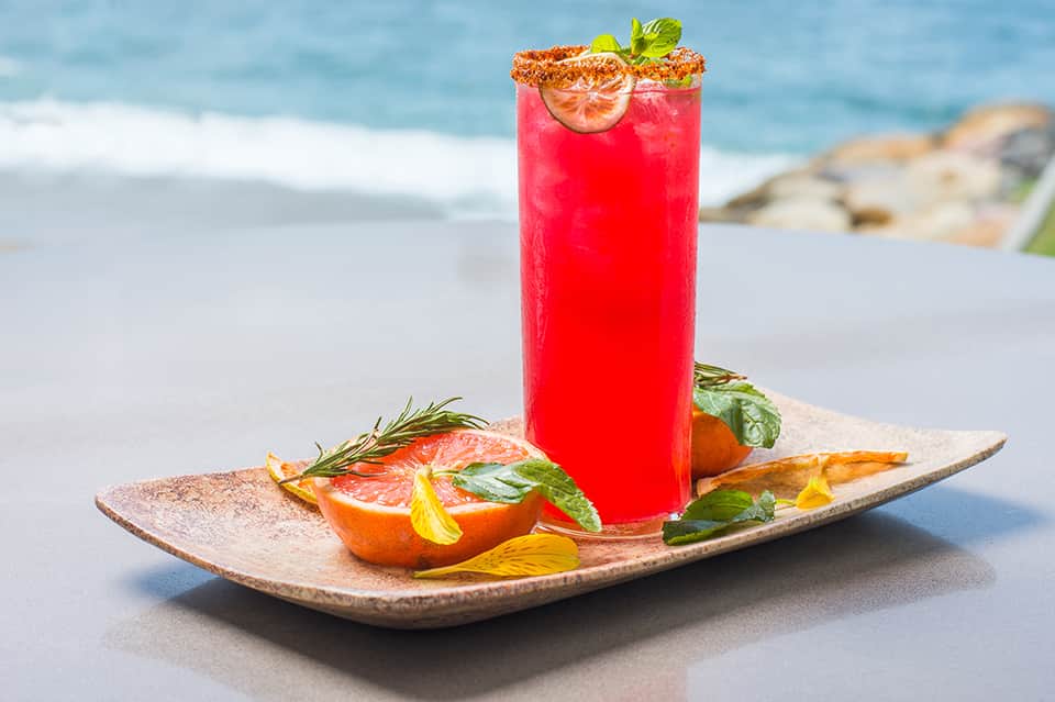 Vibrant red cocktail with grapefruit slice and spice rim on beach with ocean backdrop