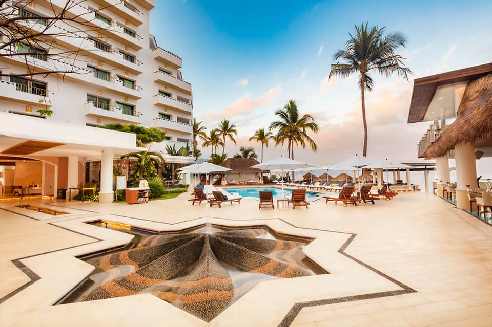 Beachfront resort pool area with palm trees, loungers, umbrellas, and modern architecture