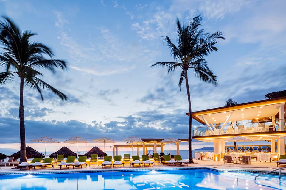 Luxury beachfront resort pool at dusk with palm trees, lounge chairs, and ocean views