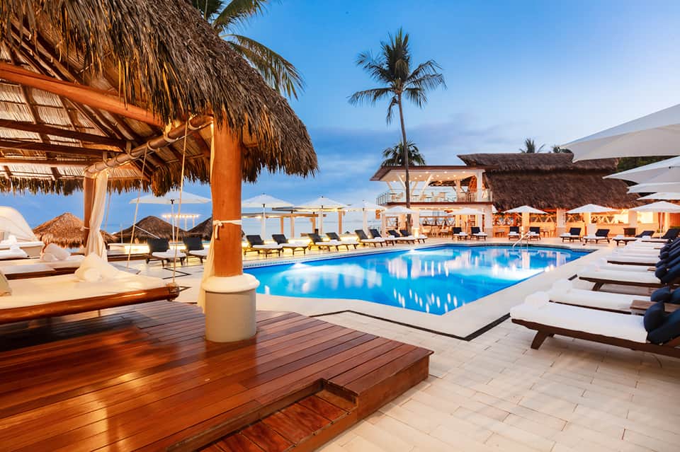 Luxury resort pool at dusk with thatched umbrellas, wooden deck, lounge seating, and palm trees