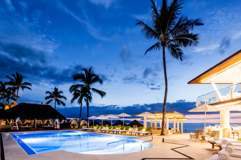 Illuminated resort pool at twilight with palm trees and ocean view in background