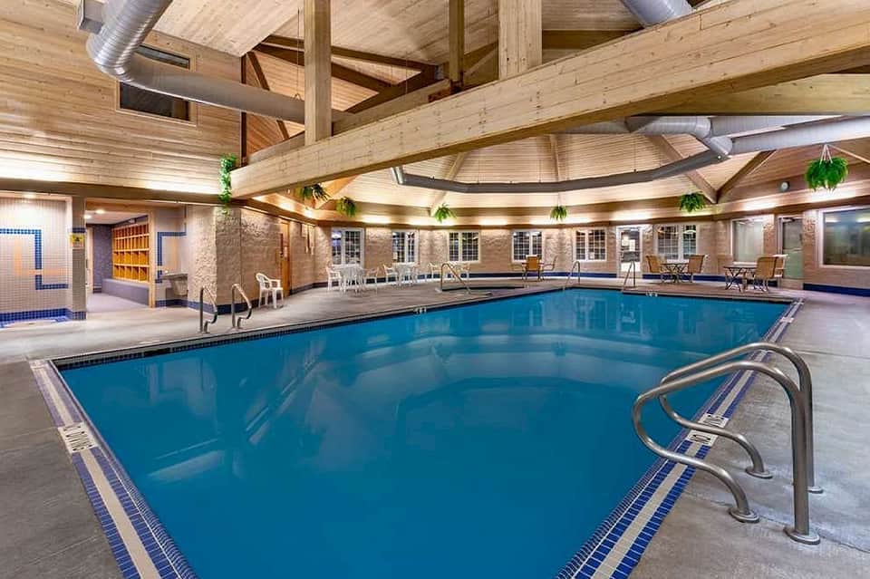 Indoor heated pool with wooden beam ceiling, seating areas, and blue water