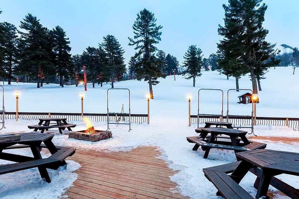 Winter resort grounds with snow-covered landscape, fire pits, picnic tables, and pine trees at dusk