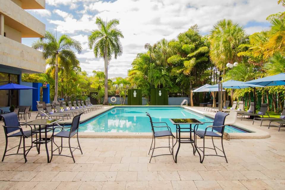 Resort pool surrounded by palm trees, lounge chairs, and blue umbrellas under cloudy sky
