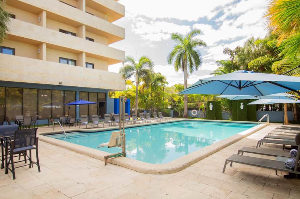 Resort pool area with turquoise water, blue umbrellas, lounge chairs, palm trees, and modern building