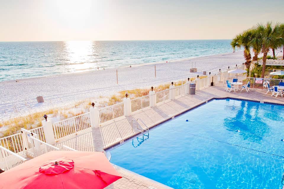 Beachfront resort pool with pink umbrellas, white fencing, and palm trees overlooking the ocean