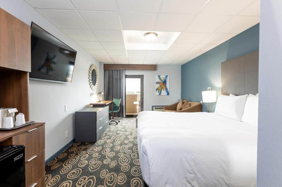 Modern hotel room with queen bed, kitchenette, TV, and contemporary decor in blue tones
