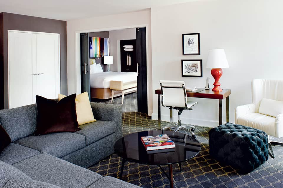 Modern hotel suite living room with gray sofa, desk, and view to bedroom