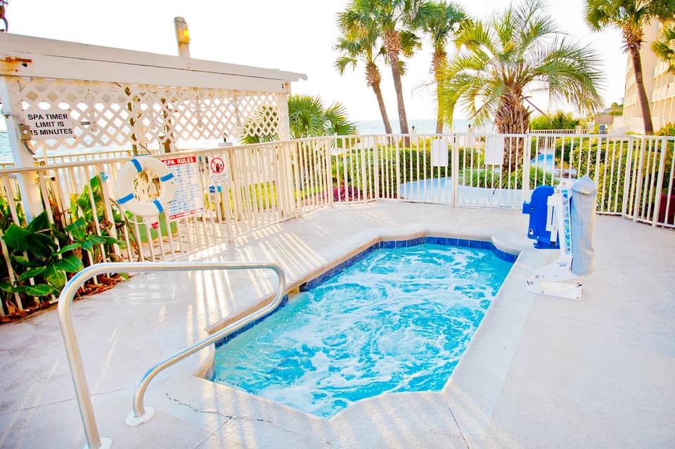 Small rectangular pool surrounded by white fencing with palm trees and beachfront buildings in background