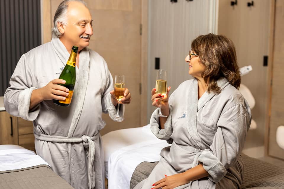 Couple in spa robes toasting with champagne in hotel room with bed and modern furnishings