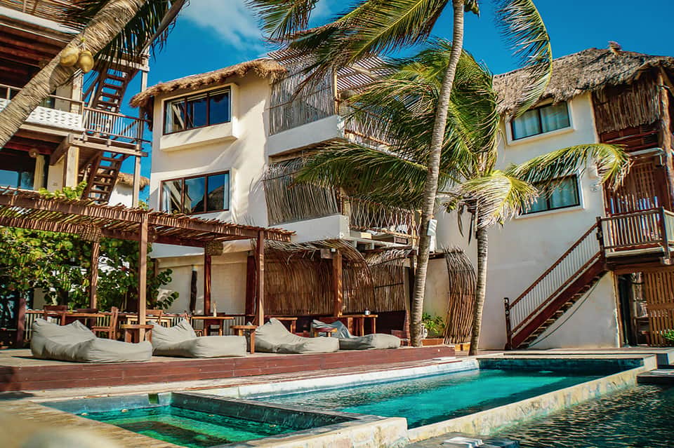 Beachfront resort buildings with thatched-roof structures, white walls, turquoise pool, and palm trees