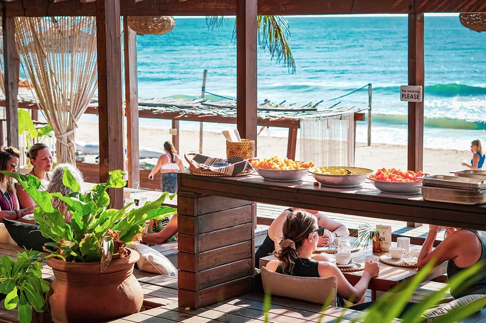 Beachfront restaurant pavilion with diners, wooden counters with food, and ocean views