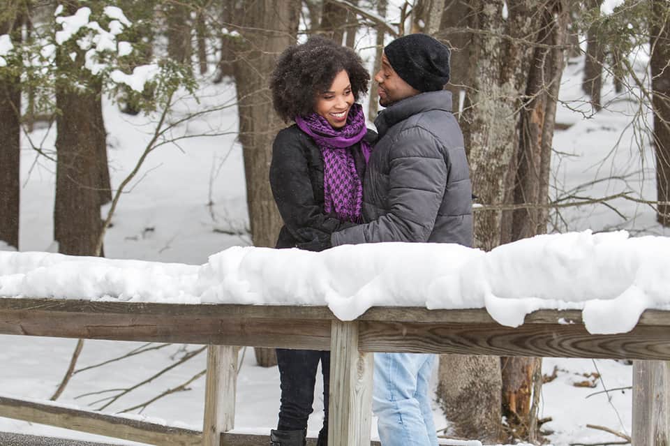 Couple in winter clothing embracing on snowy wooden fence with frozen lake and bare trees behind