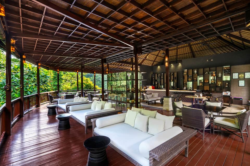 Indoor-outdoor lounge with woven seating, wooden deck, tropical plants, and natural lighting