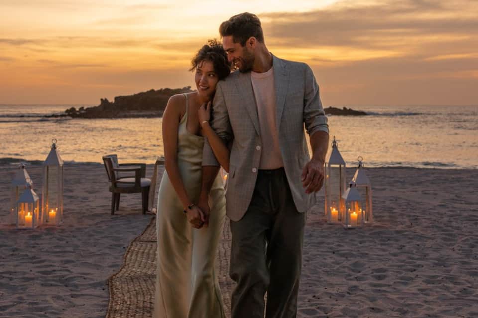 Couple walking on beach at sunset with lit lanterns and chairs in background