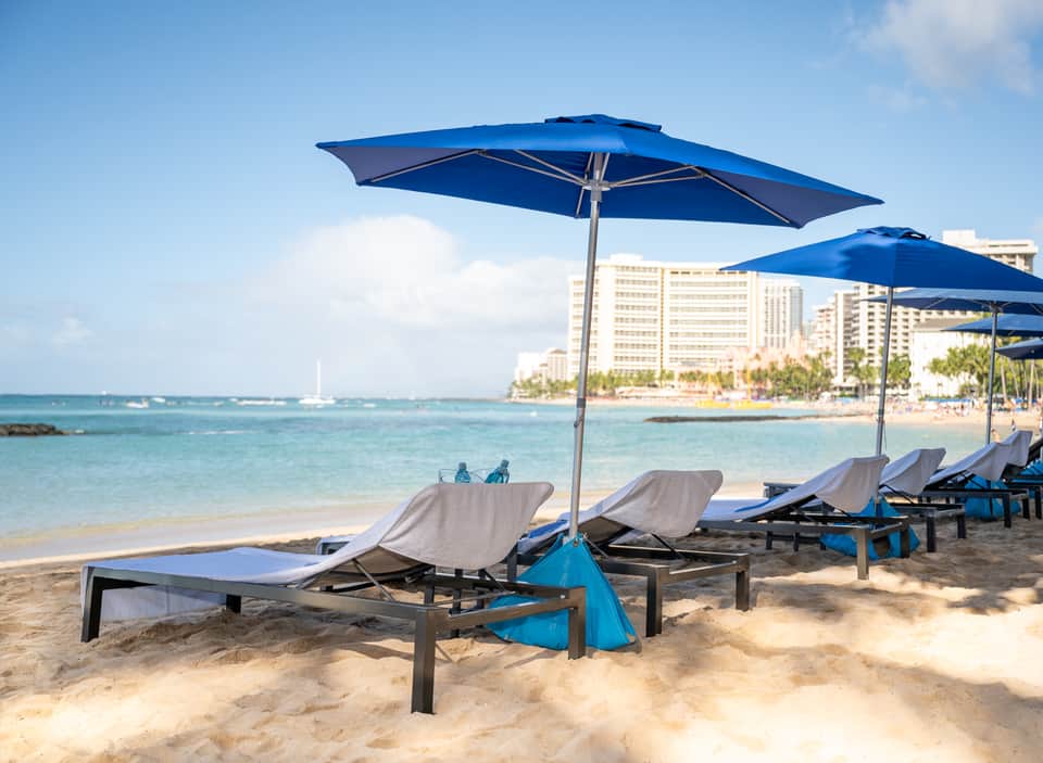 Beach lounge chairs with blue umbrellas on sandy shore with turquoise ocean and resort buildings