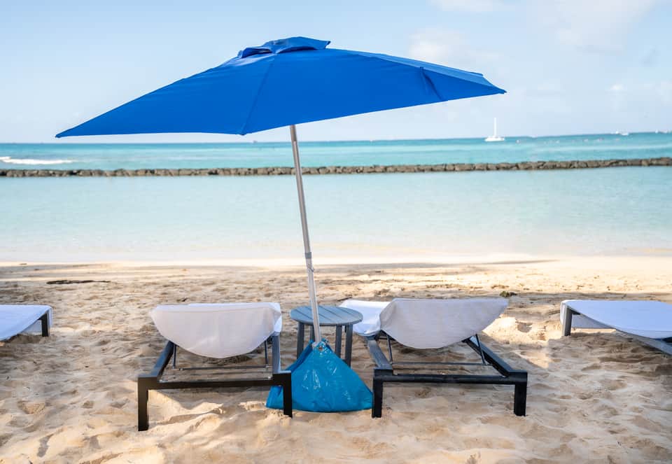 Two beach lounge chairs with white cushions under blue umbrella facing calm turquoise ocean