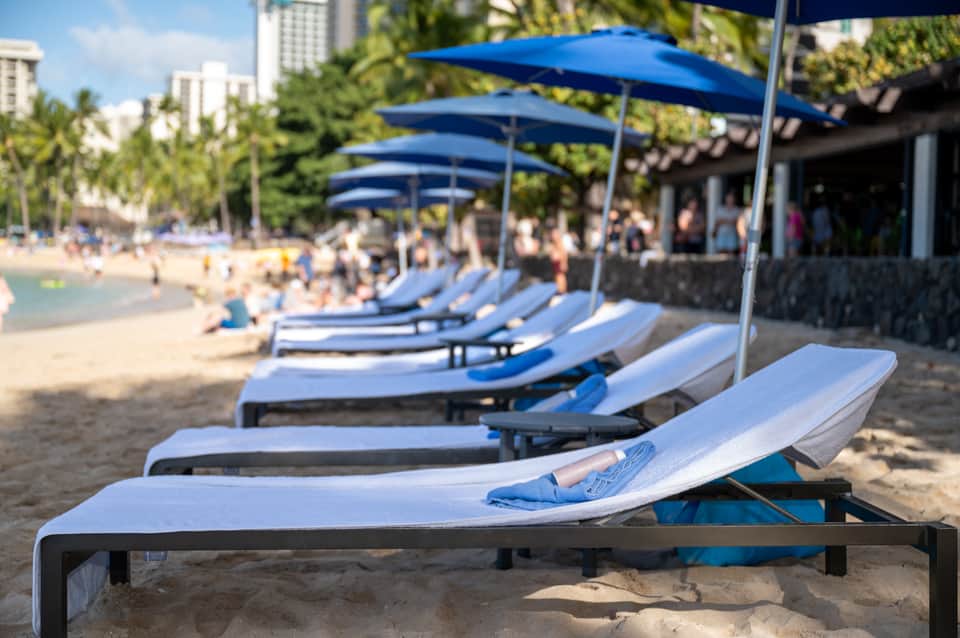 Row of white and blue lounge chairs with umbrellas on sandy beach near ocean and palm trees