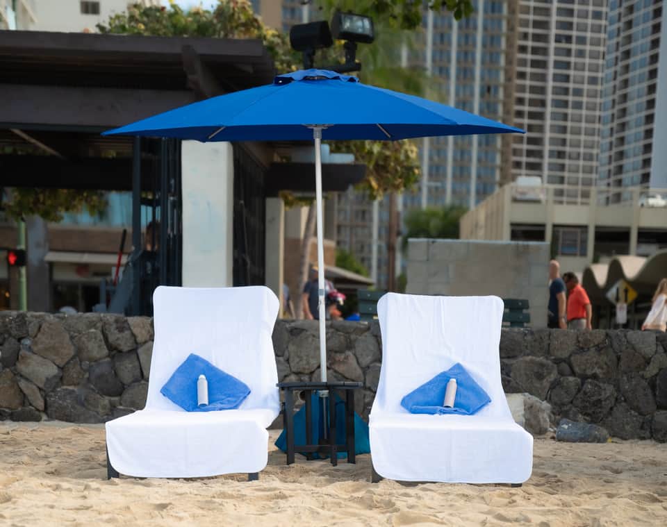 Two white pool loungers under blue umbrella on urban beach with city skyline