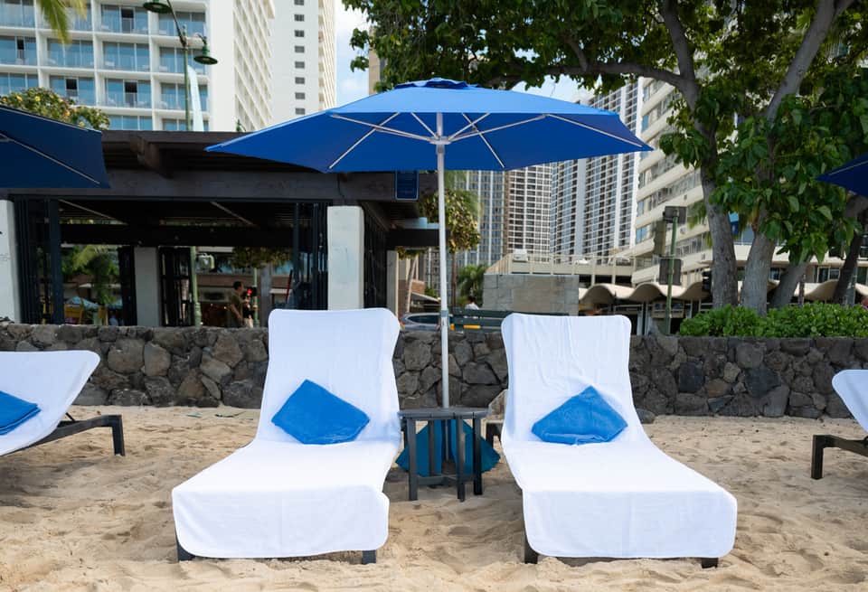 Two lounge chairs with blue pillows and umbrella on sandy beach area