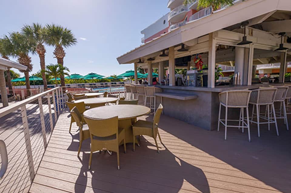 Waterfront deck with yellow chairs, tables, bar, and pool umbrellas near palm trees