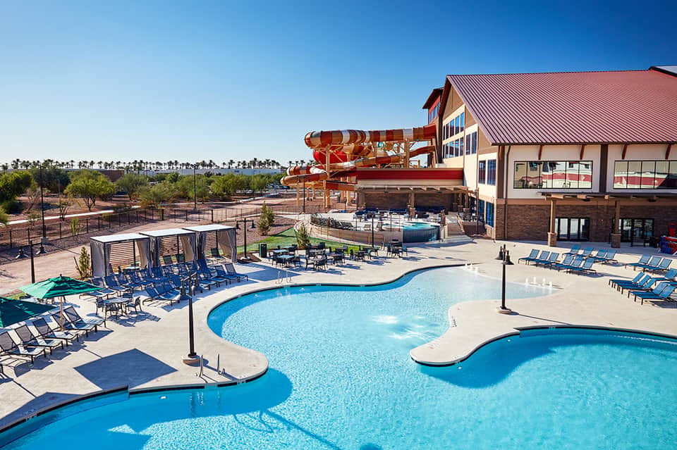 Resort pool area with water slide, lounge chairs, and facility building under blue sky