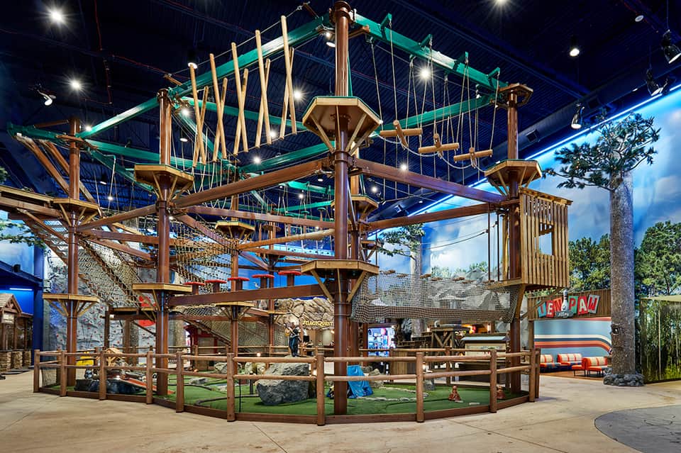 Indoor adventure playground with wooden ropes course, climbing structures, green elements, and blue sky ceiling murals