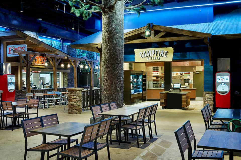 Indoor entertainment venue with campfire-themed seating areas, wooden structures, red vintage appliances, and blue ceiling