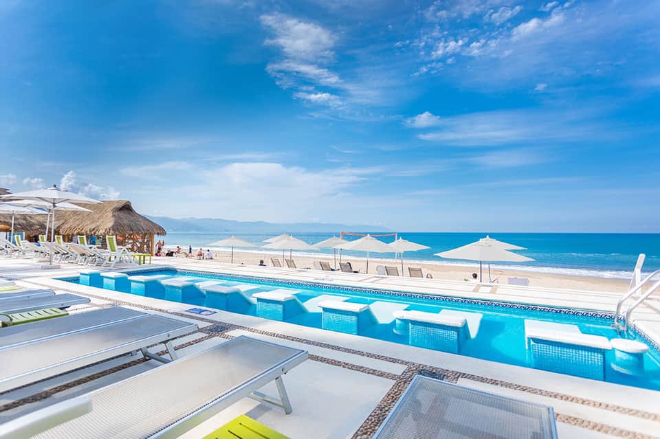 Bright blue resort pool with geometric white sun loungers overlooking pristine beach and turquoise ocean