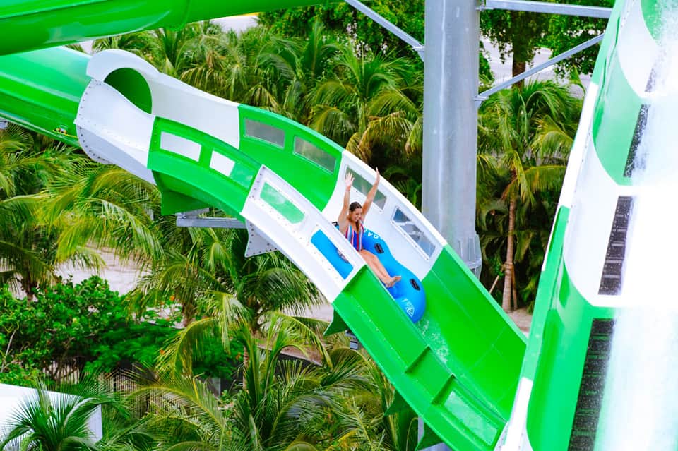 Person sliding down a bright green water slide at a tropical resort surrounded by palm trees
