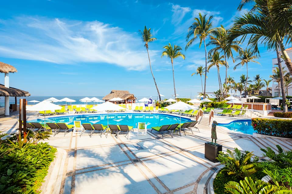 Resort pool with bright blue water, yellow loungers, palm trees, and beachfront view
