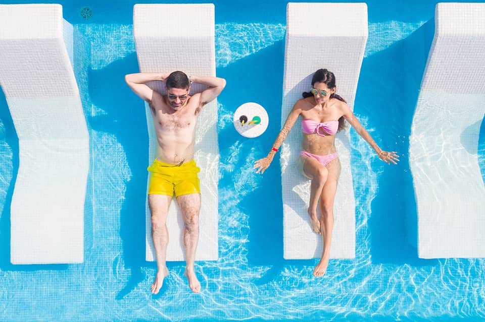 Two people floating on white loungers in bright blue pool with mirrored reflections below