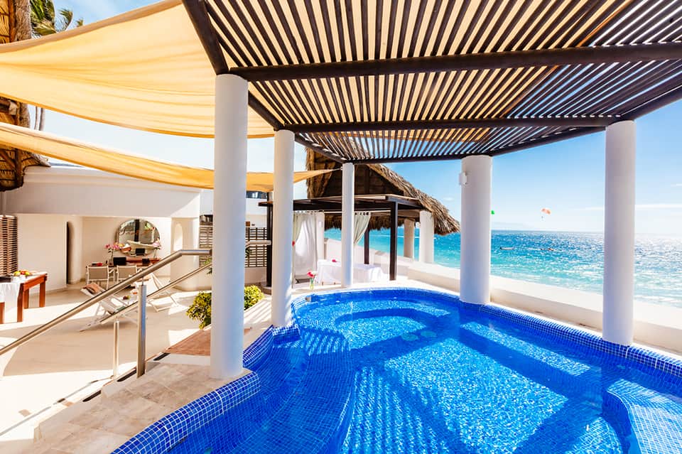 Infinity pool with blue tiles overlooking turquoise ocean, white pergola, and beach cabanas