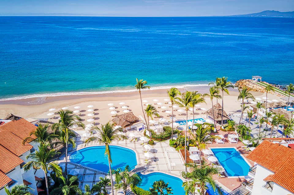 Beachfront resort with multiple pools, palm trees, white umbrellas, and ocean view