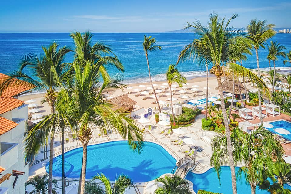 Aerial view of beachfront resort with pool, palm trees, sandy beach, and ocean horizon