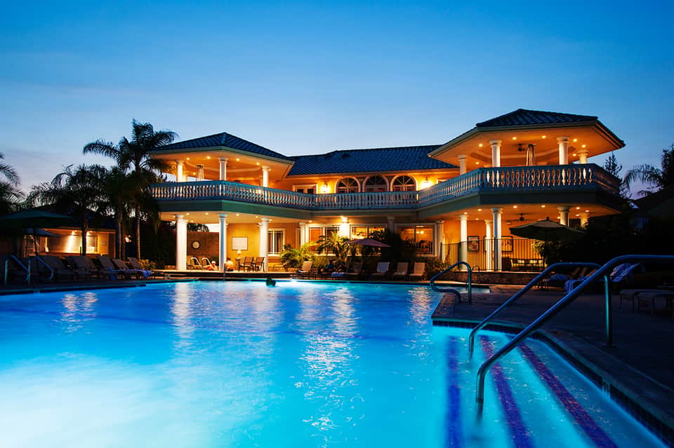 Illuminated two-story luxury home with orange-lit exterior, wraparound balconies, blue swimming pool, and lounge chairs at dusk