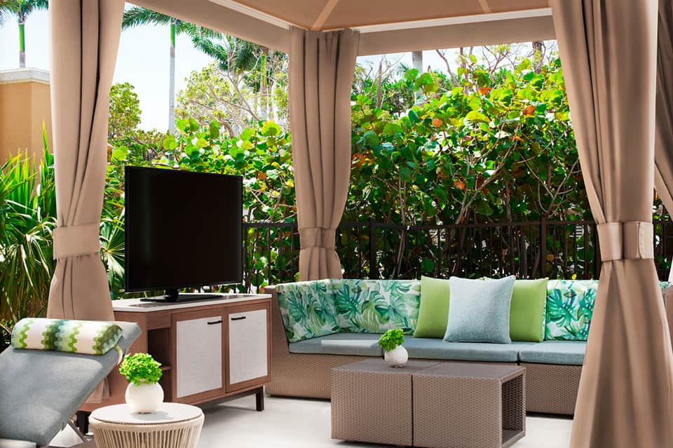 Covered patio with wicker furniture, green cushions, TV, white curtains, and tropical vegetation views