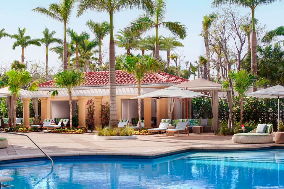 Resort pool with cabanas, red-roofed building, palm trees, and lounge seating areas