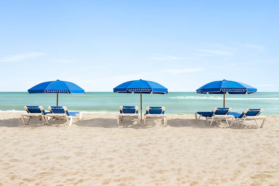 Beach scene with three blue umbrellas, white lounge chairs, turquoise ocean, and sandy shore