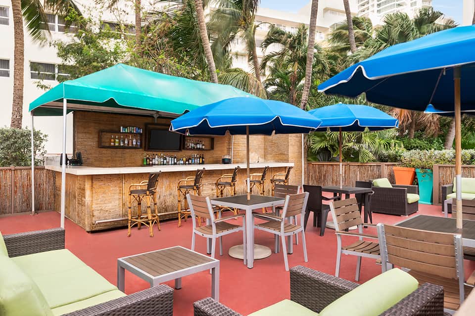 Beachside bar with turquoise umbrellas, wooden counter, bar seating, and tropical palm trees
