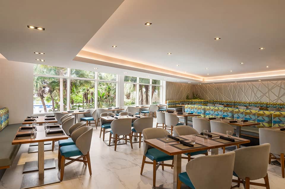Contemporary restaurant with large windows, palm tree views, wooden tables, and beige upholstered chairs