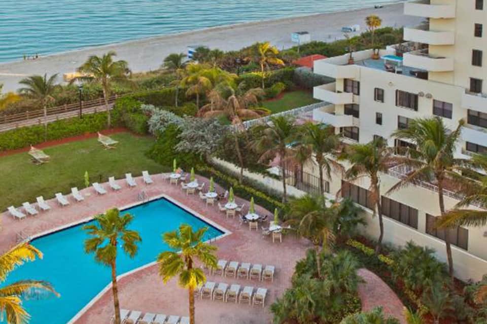 Aerial view of resort pool, lawn, and white apartment buildings overlooking beach