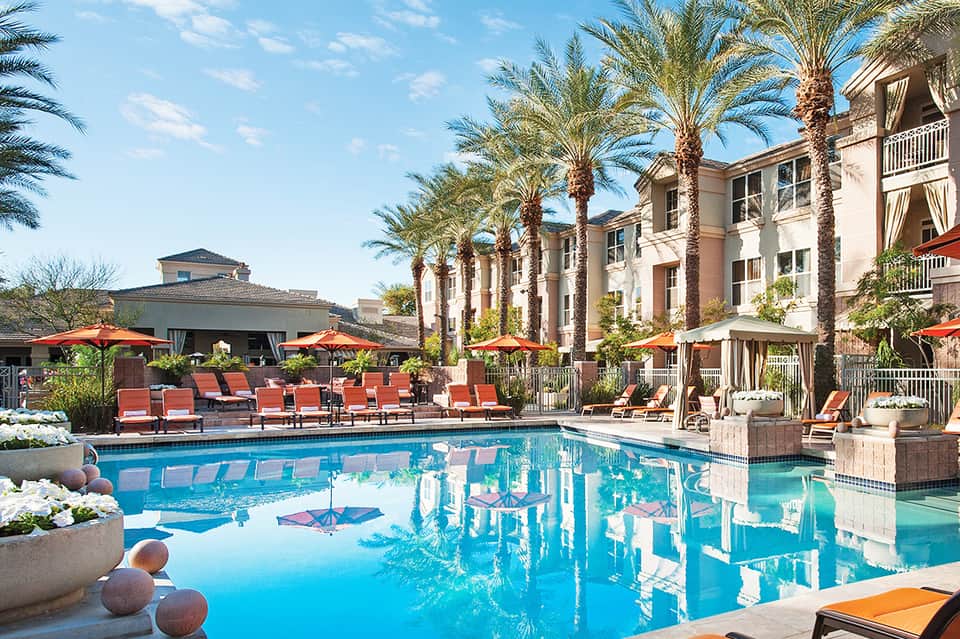 Resort pool area with blue water, red lounge chairs, palm trees, and building facade