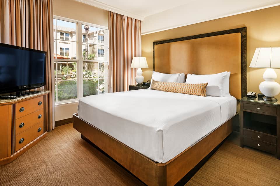 Hotel bedroom with gold headboard, warm lighting, wood furnishings, and garden view