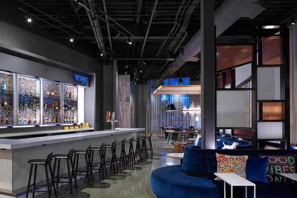 Modern hotel bar and lounge with blue accent seating, illuminated shelving, and industrial ceiling
